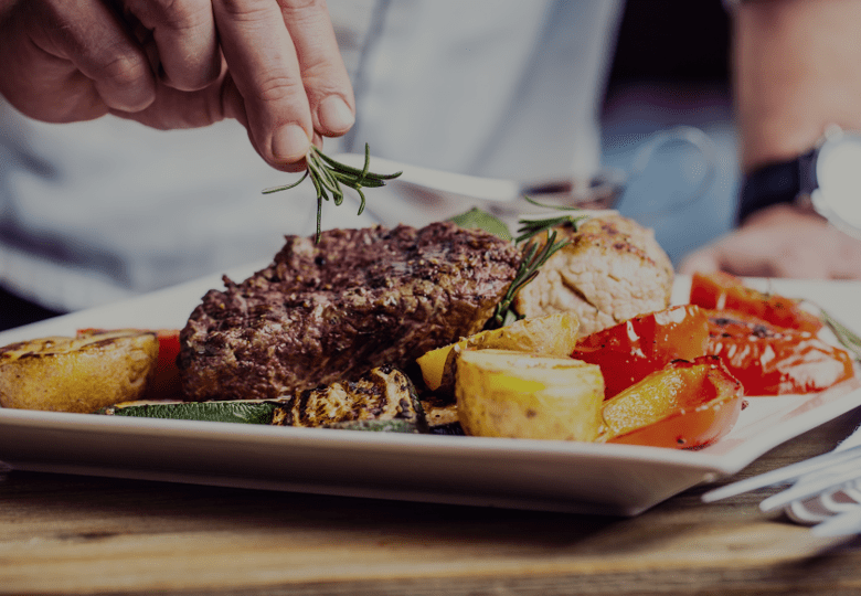 Chef garnishing grilled steak with herbs beside roasted vegetables